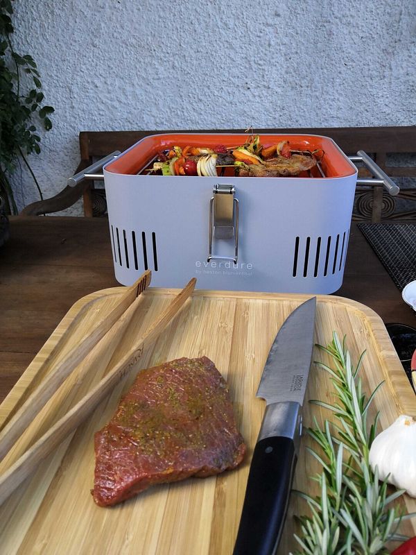 Auf dem Schneidebrett vor dem Grill liegt das nächste Stück Fleisch bereit, der Grillrost ist noch voll