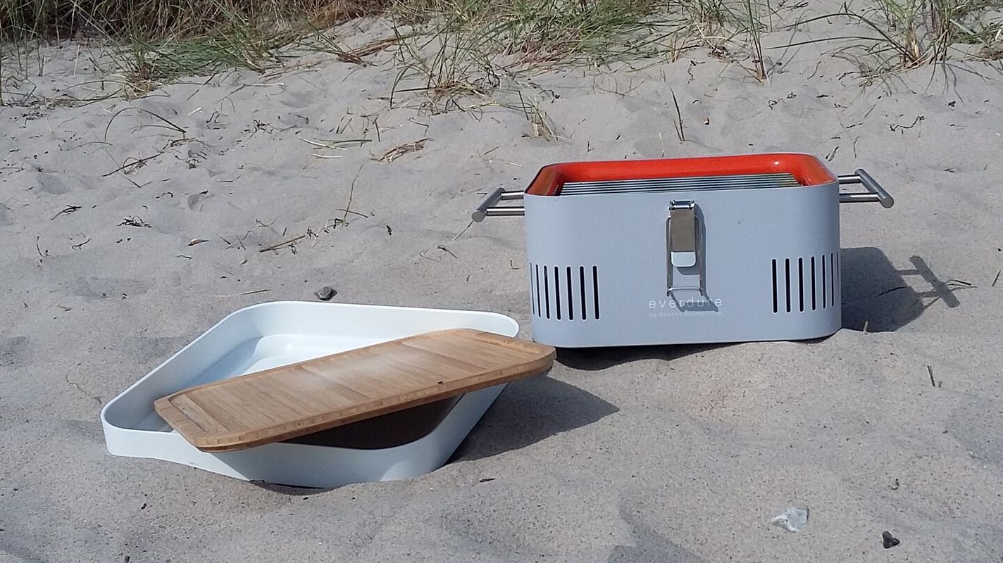 Cube Grill steht vor einer Düne am Strand, die weiße Aufbewahrungsbox und das Schneidebrett aus Bambus liegen daneben