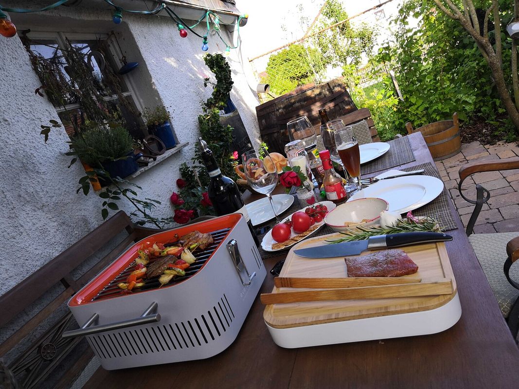 Gedeckter Gartentisch aus Holz, Fleisch, Messer, Grillzange und Rosmarin  liegen auf dem Schneidebrett, der Everdure Cube steht daneben, auf dem Grillrost drei Gemüsespiese und zwei Rinderfilets
